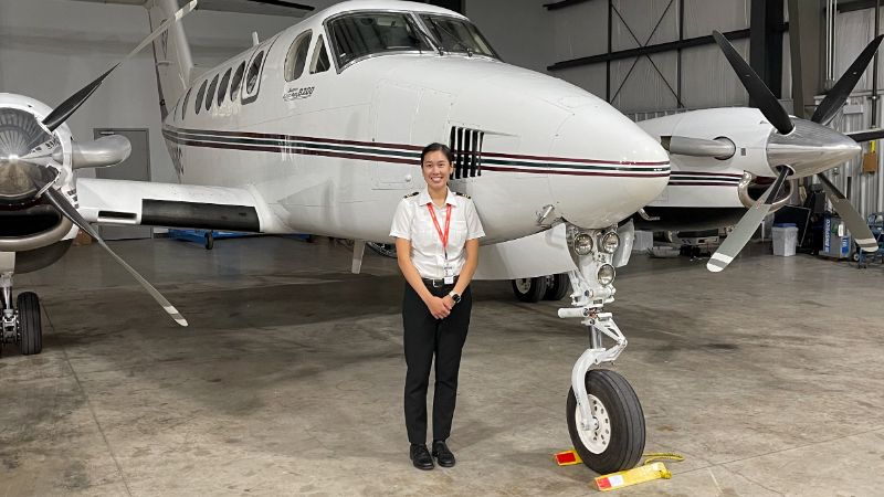 A person standing in front of an aircraft inside a hangar, wearing a pilot uniform and smiling at the camera.