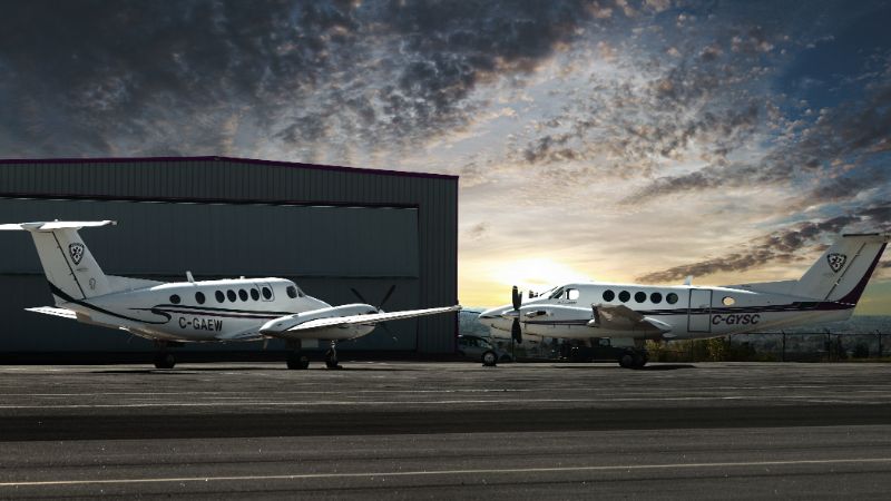 Image shows two aircraft parked side by side in front of a hangar, with dramatic sky at sunset in background.