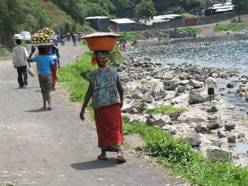 A woman carrying a large basket on her head, walking along a path by a rocky riverside. She is wearing a colorful top and a red skirt. Other people are walking behind her, also carrying items.