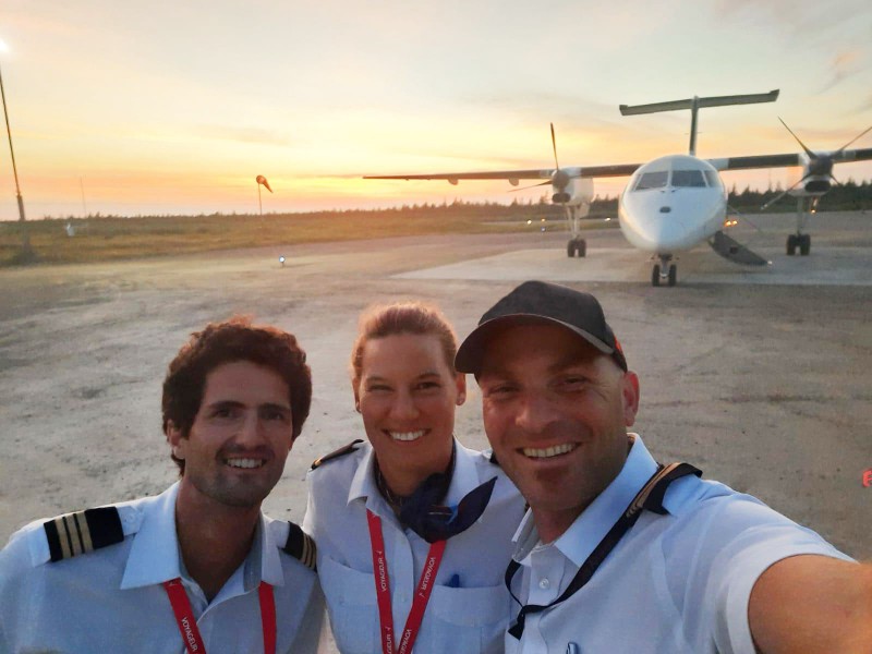 Three smiling pilots, two men and one woman, taking a selfie in front of an airplane at sunset. They are standing on the tarmac, and the aircraft is visible in the background.