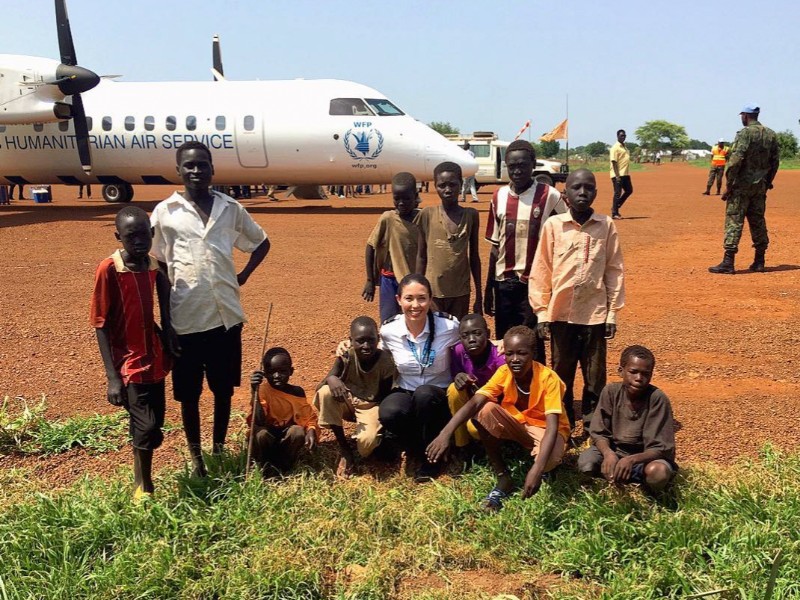 A group of children and a woman sitting and standing together in front of an aircraft labeled Humanitarian Air Service. The group is posing for a picture on a dirt airstrip.