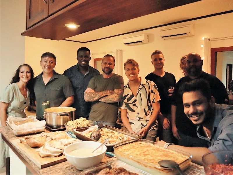 A group of people smiling and standing together around a table filled with food dishes inside a dining area. They are enjoying a meal together.