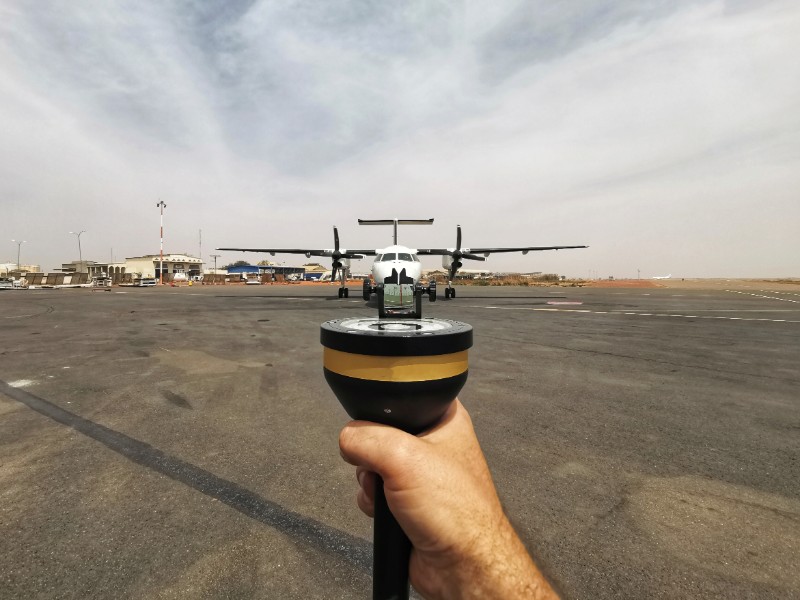 A hand holding a measuring device pointed towards an airplane on a tarmac in a dusty environment. The background shows airport buildings and equipment.