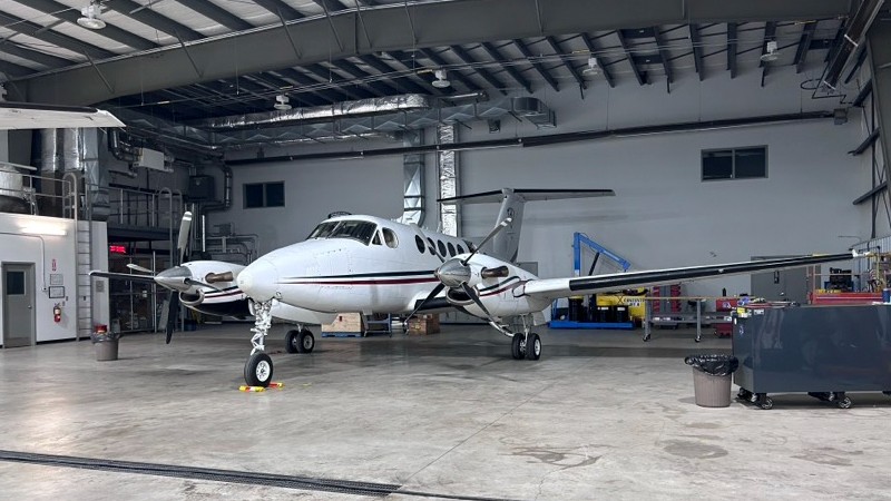 An aircraft inside a spacious hangar, with various tools and equipment visible in the background. 