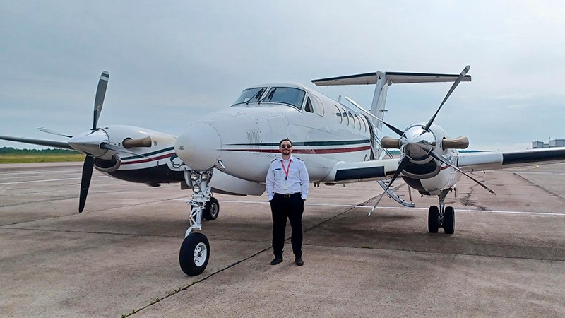 A person in a pilot uniform standing in front an aircraft on the tarmac, smiling and posing for the camera.