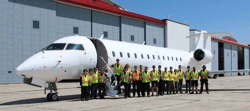 A group of people wearing high-visibility vests standing in front of a parked aircraft on the tarmac. The backdrop includes a Voyageur Aviation hangar with a red roof under a clear blue sky.