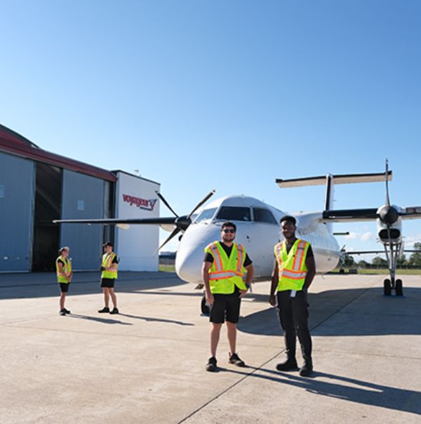 Four people wearing high-visibility vests standing on a tarmac in front of a large aircraft. The background shows an open hangar with the Voyageur Aviation logo.