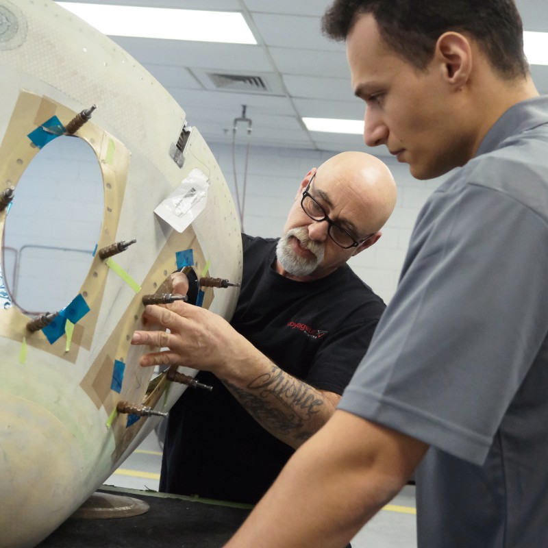 Two technicians working on an aircraft component in a workshop. One technician, with glasses and a beard, is pointing at the component while explaining something to the other technician, who is attentively watching. The component has markings and tape indicating areas of work. 