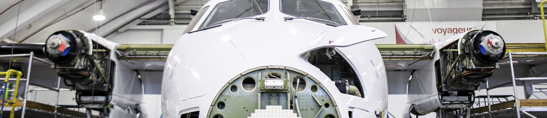 A front view of an aircraft inside a large hangar. The aircraft's nose is prominent, with its engines partially disassembled and exposed on either side. The hangar has overhead cranes and various equipment around the aircraft.